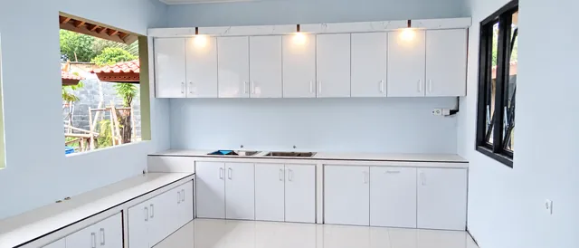 Kitchen set putih bentuk L dengan lampu sorot di bawah kabinet atas dan jendela di samping